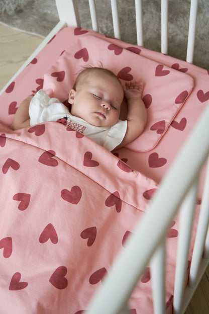 Baby wrapped in pink bedding with heart design, photographed through crib rails