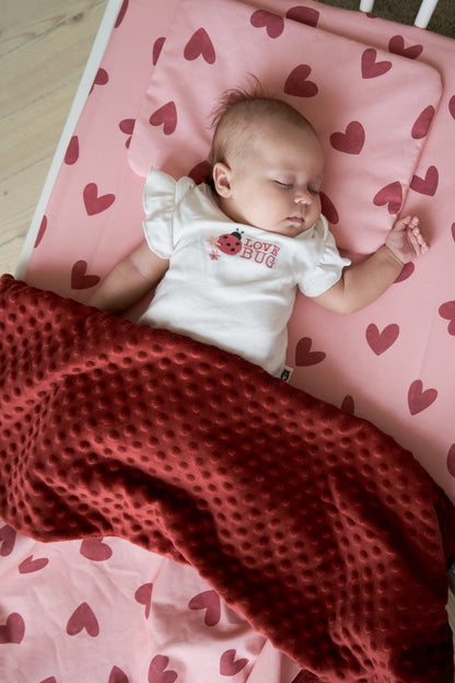 Sleeping baby on the Heartland bedding set, with red heart prints on a pink sheet and a maroon minky blanket, creating a cozy and adorable scene